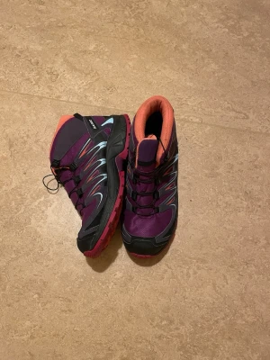 Lila och svarta kängor från Salomon - Säljer ett par sportiga kängor från Salomon i lila och svart med röda detaljer och orange insida. Skorna har snörning och robust platt sula, perfekta för utomhusaktiviteter. Materialet är syntet och skinn, designen är modern och funktionell.