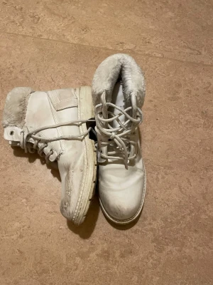 Vita boots med fuskpäls - Säljer ett par vita boots med snörning och mjuk fuskpäls runt skaftet. Skorna har en platt sula och rund tå, perfekta för kalla dagar. Materialet är syntet med pälsdetaljer som ger extra värme och stil.
