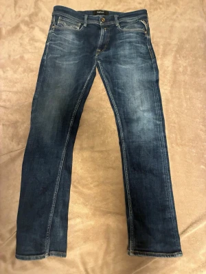 Blå Replay jeans straight fit - Snygga blå jeans från Replay med klassisk femficksdesign och slitna detaljer. Modellen har raka ben och normal passform, med Replay-logga på bakfickan och läderpatch i midjan. Perfekt för dig som gillar en tidlös jeanslook.
