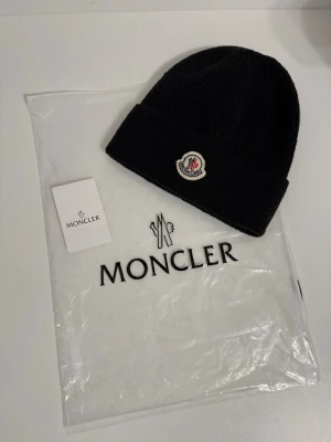Svart Moncler mössa i ull - Svart ribbstickad mössa från Moncler med klassisk uppvikt kant och ikonisk Moncler-logga framtill. Tillverkad i mjuk ull som håller dig varm och snygg under kyliga dagar. Perfekt accessoar för dig som gillar stilrena detaljer och exklusiva märken.