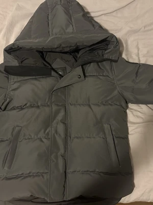 Mörkgrå pufferjacka från Canada Goose Black label - Säljer en mörkgrå pufferjacka från Canada Goose med huva och snyggt broderat logomärke på ärmen. Jackan har dragkedja, tryckknappar och två sidofickor. Perfekt för kalla dagar med sin quiltade design och rejäla passform.