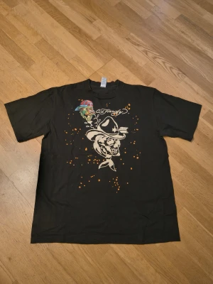 Svart Ed Hardy t-shirt med tryck - Cool svart t-shirt från Ed Hardy med stort tryck av döskalle och hatt framtill samt färgglada detaljer och logga både fram och bak. T-shirten har korta ärmar och är i mjuk bomull, perfekt för en streetwear-look.