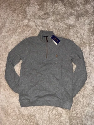 Grå half zip tröja från Polo Ralph Lauren - Snygg grå långärmad tröja från Polo Ralph Lauren med half zip och klassisk krage. Tröjan har det ikoniska broderade Polo-logot på bröstet och är tillverkad i mjuk bomull. Perfekt för chill dagar och enkel att matcha med jeans eller joggers.