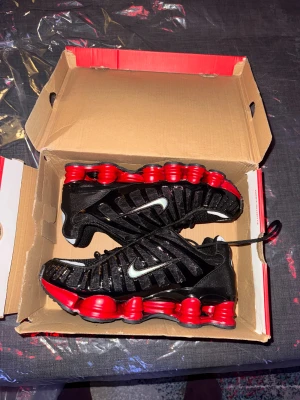 Nike Shox sneakers svart/röd - Nike Shox sneakers med svart mesh och glansiga detaljer, samt en ikonisk röd Shox-plattformssula. Swoosh-logga i silver på sidan och snörning framtill. Skorna har en sportig och futuristisk vibe, perfekta för dig som gillar att sticka ut.