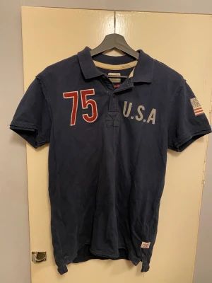 Mörkblå Vintage Co  - Snygg mörkblå t-shirt från Jack & Jones Vintage Co med tryck '75' och 'U.S.A' på framsidan samt stort 'Vintage Co 75' på ryggen. Amerikansk flagga på ärmen och klassisk krage. Tillverkad i mjuk bomull, perfekt för en avslappnad stil.