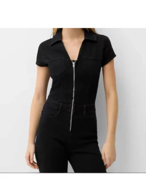 Svart jumpsuit med dragkedja - Snygg svart jumpsuit med korta ärmar, krage och markerad midja. Framtill finns en silverfärgad dragkedja hela vägen ner och två bröstfickor. Byxbenen är vida för en cool och avslappnad look. Perfekt för dig som vill sticka ut.