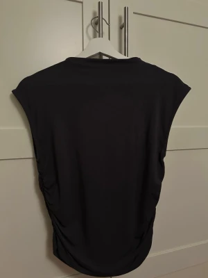 Svart croppad topp med rynkade sidor strl. L - Snygg svart croppad topp med höga axlar och rynkade sidor. Toppen är ärmlös och har en tight passform som framhäver figuren. Färgen är mörkgrå.