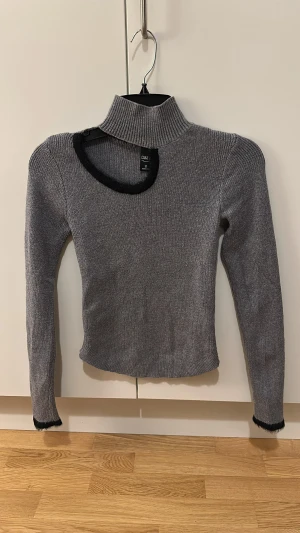 Grå stickad polotröja från Dazy - Säljer en grå stickad polotröja från Dazy med svart kant vid halsringningen och ärmsluten. Tröjan har lång ärm, ribbad struktur och en cool asymmetrisk cut-out vid halsen som ger en unik look.