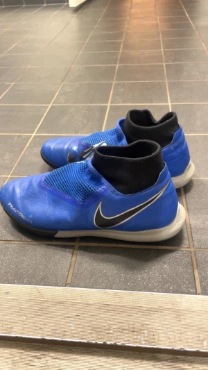 Blå Nike Phantom Vision fotbollsskor - Säljer ett par blå Nike Phantom Vision fotbollsskor (innomhusskor) med svart och vit swoosh på sidan. Skorna har en strumpliknande svart krage och är tillverkade i syntetmaterial med meshdetaljer för extra ventilation. Snygg och modern design som sticker ut på planen.  Pris kan diskuteras..
