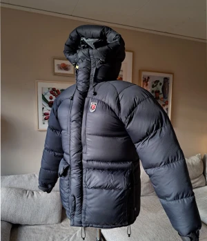 Mörkblå  dunjacka från Fjällräven - Säljer en mörkblå , riktigt fet dunjacka från Fjällräven med stor huva och klassisk röd logga på bröstet. Jackan har dragkedja, snörning och två stora fickor framtill. Perfekt för kalla vinterdagar när du vill hålla dig varm och ändå se chill ut.