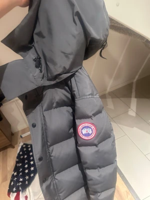 Grå dunjacka från Canada Goose - Säljer en grå dunjacka från Canada Goose med huva och klassisk logga på ärmen. Jackan har dragkedja och tryckknappar framtill, samt justerbar huva med snörning. Perfekt för kalla vinterdagar och har en clean, modern look.