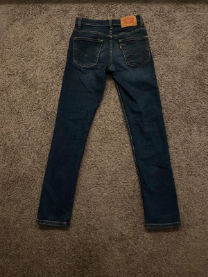 Levi's 512 Slim Taper jeans mörkblå 146/152 - Snygga Levi's 512 Slim Taper jeans i mörkblå tvätt med klassiska fem fickor och gul kontrastsöm. Storlek 146/152. Modellen har smal passform med avsmalnande ben och normal midja. Perfekt för dig som gillar stilrena och moderna jeans.