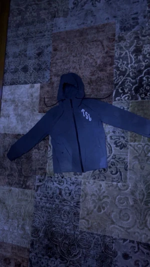 Svart vindjacka Trapstar Shooters - Svart vindjacka med huva och dragkedja framtill. På bröstet står det 'TSU' och på ryggen finns en stor logga med pantermotiv och texten 'Tripstar Shooters'. Jackan har långa ärmar och praktiska fickor med dragkedja.