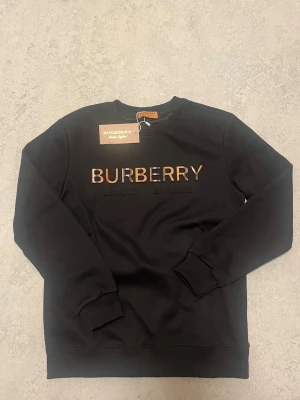 Svart sweatshirt från Burberry - Svart sweatshirt från Burberry med klassisk logga i brunt och beige framtill. Tröjan har rund halsringning, långa ärmar och ribbade muddar vid ärmslut och nederkant. Perfekt för en stilren och trendig look.