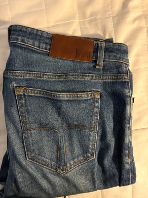 Blå jeans från Tiger of Sweden - Snygga blå jeans från Tiger of Sweden med klassisk femficksdesign och brunt läderpatch bak i midjan. Jeansen har raka ben och subtila slitningar framtill för en cool look. Passar dig som gillar stilrena och tidlösa jeans.