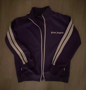 Lila Palm Angels zip hoodie - Säljer en lila hoodie från Palm Angels med vita och svarta ränder längs ärmarna. Tröjan har dragkedja framtill, hög krage och logga på bröstet. Perfekt för dig som gillar streetwear och vill sticka ut lite extra.