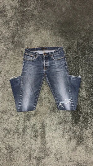 Blå slim fit jeans från Nudie - Snygga blå slim fit jeans från Nudie med slitningar och ett stilrent hål på ena knät. Jeansen har klassisk femficksdesign, fransade benslut och den ikoniska Nudie-loggan på bakfickan. Perfekt tvättad look och en slim fit-passform som sitter riktigt bra längs benen.  Har du frågor är det bara att skicka! Priset är inte hugget i sten – kom med ett rimligt bud.