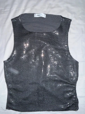 Glittrig svart croppad topp FB Sister - Säljer en glittrig svart croppad topp från FB Sister i storlek S. Toppen är ärmlös och har ett mesh-liknande material med små paljetter som ger en snygg glans. Baktill finns en knytning för extra cool passform. Perfekt för att sticka ut med lite extra bling.