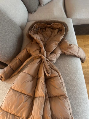 Lång brun pufferjacka med fuskpäls - Snygg lång pufferjacka i brun nyans med huva och fluffig fuskpäls runt kanten. Jackan har bälte i midjan helt splittrans ny endast testad men Tyvär så köpte jag fel storlek och jag orkar inte returnera 