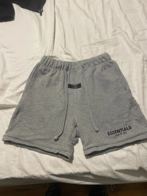 Grå Essentials Fear of God shorts - Säljer ett par grå mjukisshorts från Essentials Fear of God i storlek S. De har elastisk midja med snörning, två sidofickor och logga tryckt på benet. Materialet är mjuk bomull som är perfekt för chill eller streetwear. Klassisk loose fit och enkel design.