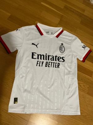 AC Milan Tröja - AC Milan bortatröja i vitt med svarta och röda detaljer från Puma. Tröjan har klubbmärke och Puma-logga på bröstet samt Emirates Fly Better-tryck. Ränder i tyget och ribbade muddar med röd och svart kant. Perfekt för fotbollsfans.