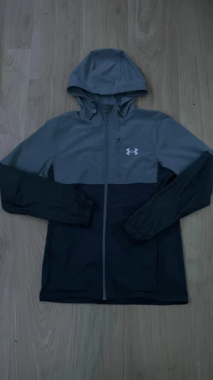 Grå och svart vindjacka Under Armour - Snygg tvåfärgad vindjacka från Under Armour i grått och svart. Jackan har huva, hel dragkedja framtill, lång ärm och en bröstficka med dragkedja. Perfekt för dig som gillar sportig stil och vill ha något lätt och funktionellt.