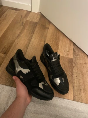 Valentino Rockrunner sneakers svart/silver - Snygga Valentino Rockrunner sneakers i svart och silver med coolt kamouflagemönster. Skorna har snörning, platt sula och detaljer i skinn och mocka. Ikoniska nitar baktill och rund tå ger en unik vibe. Perfekta för dig som vill sticka ut med stil.