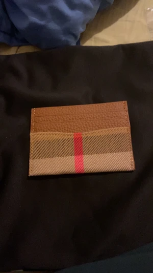 Rutig plånbok från Burberry - Snygg och stilren plånbok från Burberry i klassiskt rutigt mönster med en röd rand i mitten. Plånboken är brun med detaljer i skinn och har flera fack för kort. Perfekt för dig som gillar lyxiga accessoarer med ikonisk design.