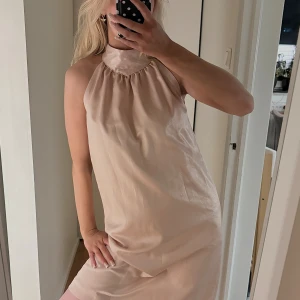 Champagnefärgad ärmlös klänning - Säljer en stilren champagnefärgad klänning med hög hals och ärmlös design. Klänningen har en rak, loose passform och är tillverkad i ett glansigt material som ger en lyxig känsla. Perfekt för dig som gillar minimalistisk och chic stil. Vet ej märket men känns som zara klänninh