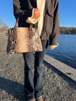 Handväska med ormmönster och bruna detaljer - Snygg handväska med ormmönster i beige och bruna toner. Väskan har två stadiga handtag i brunt och en praktisk dragkedja på baksidan. Perfekt storlek för att få plats med allt du behöver och ger en cool touch till din outfit.