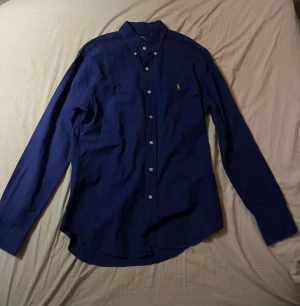 Blå skjorta Polo Ralph Lauren M - Klassisk blå skjorta från Polo Ralph Lauren i slim fit med button down-krage och broderad gul logga på bröstet. Skjortan har långa ärmar och vita knappar framtill. Perfekt för dig som gillar stilren och tidlös design. Skriv om ni har några frågor