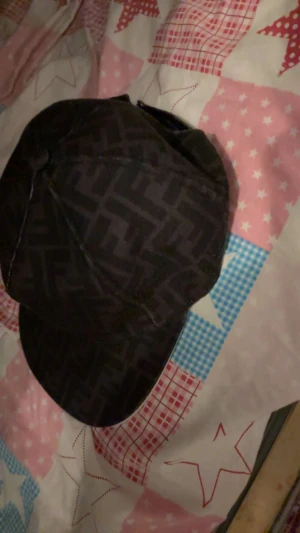 Svart Fendi keps med FF-mönster - Snygg svart keps från Fendi med det klassiska FF-logomönstret över hela ytan. Kepsen har böjd skärm och justerbar rem baktill. Tillverkad i ett slitstarkt tygmaterial som känns lyxigt och trendigt. Perfekt för dig som gillar designeraccessoarer.