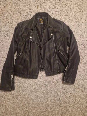 Svart skinnjacka från Mywear Woman XS - Snygg svart skinnjacka från Mywear Woman i storlek XS. Jackan har klassisk bikerstil med silvriga dragkedjor, bred krage och detaljerade sömmar på axlar och ärmar. Perfekt för dig som vill ha en edgy look.