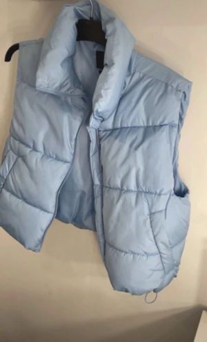 Ljusblå quiltad dunväst (endast testad)  - Snygg ljusblå dunväst med quiltad design och hög krage. Västen har dragkedja framtill och är perfekt för lager-på-lager-stil. Den har en modern, lite oversized passform och är tillverkad i syntetmaterial som håller dig varm under kyliga dagar.