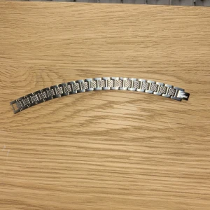 Silverfärgat Lacoste armband med mönster - Stilrent lacoste armband i silverfärgad metall med rektangulära länkar. Varje länk har ett graverat rutmönster och en av länkarna har en ingraverad logotyp. Perfekt accessoar för att lyfta din outfit med en cool och modern touch.