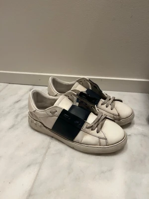 Valentino Open sneakers vit/svart - Snygga Valentino Open sneakers i vitt skinn med bred svart läderdetalj över snörningen. Platt sula med ikoniska nitar baktill och rund tå. Skorna har grå snören och en stilren, modern look som sticker ut.