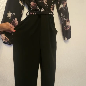 Svart byxdress med blommiga ärmar - Snygg svart byxdress med vida ben och blommiga långärmade ärmar. Byxdressen har ett matchande knytband i midjan och är tillverkad i ett mjukt, följsamt tyg. Perfekt för dig som gillar en stilren look med en touch av romantik.