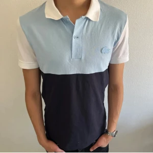 Lacoste piké t-shirt - Snygg piké från Lacoste med färgblock i ljusblått, vitt och mörkblått. Klassisk krage, korta ärmar och broderad krokodil på bröstet. Perfekt för dig som gillar stilren design och vill ha en t-shirt med lite extra edge.