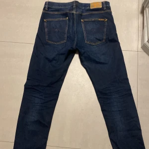 Mörkblå jeans från Nudie Jeans 32/30 - Säljer ett par mörkblå jeans från Nudie Jeans med klassisk femficksdesign och kontrastsömmar. Jeansen har raka ben och snygga detaljer på bakfickorna. Tillverkade i slitstarkt bomullstyg och passar perfekt till en avslappnad stil. Modellen är lean Dean 