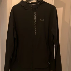 Svart vindjacka från Under Armour L - Svart vindjacka från Under Armour med huva och hel dragkedja framtill. Jackan har logga och text längs dragkedjan, samt meshfoder på insidan för extra komfort. Perfekt för träning eller när du vill ha en sportig look.