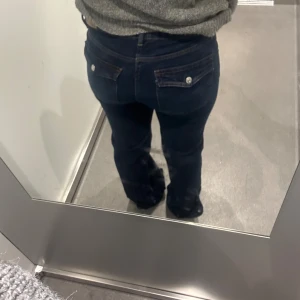Mörkblå bootcut jeans med fickdetaljer - Snygga mörkblå jeans med bootcut passform och klassiska fickor med knappdetaljer bak. Jeansen har en något utsvängd benvidd och är tillverkade i ett robust denimtyg. Perfekta för dig som gillar en tidlös och avslappnad stil.