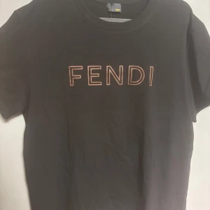 Svart Fendi t-shirt med logga - Svart t-shirt från Fendi med klassisk rund hals och korta ärmar. Framsidan pryds av Fendi-loggan i brunt tryck. T-shirten är i mjuk bomull och har en clean, stilren look som passar perfekt till jeans eller shorts.