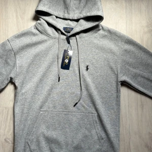 Grå hoodie från Polo Ralph Lauren - Snygg grå hoodie från Polo Ralph Lauren med klassisk broderad logga på bröstet. Tröjan har huva med snörning, känguruficka och ribbade muddar. Perfekt för en chill och stilren look. Tillverkad i mjuk bomullsmix för extra komfort.