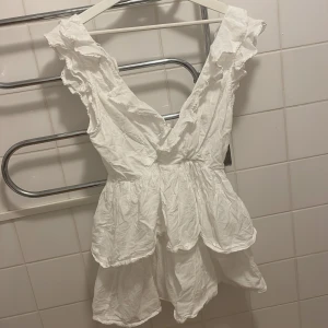 Vit volangklänning med djup rygg och knyte  - Supersöt vit kortklänning med volanger och djup v-ringad rygg. Klänningen har ärmlös design med breda axelband och omlottkänsla. Strycker självklart innan leverans 