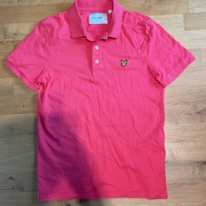 Rosa/Röd pikétröja från Lyle & Scott - Fräsch Rosa/Röd pikétröja från Lyle & Scott i ekologisk bomull. Klassisk krage, korta ärmar och knäppning med tre vita knappar. På bröstet sitter den ikoniska broderade gula örnen. Perfekt för dig som gillar stilrena och färgglada plagg. Storlek: S