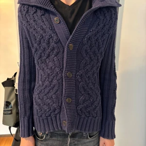 Mörkgrå stickad kofta från Hilfiger Denim - Säljer en chunky mörkgrå stickad kofta från Hilfiger Denim med grovt flätmönster, stora knappar och hög krage. Koftan har långa ärmar och ribbade muddar. Perfekt för lager-på-lager under kyliga dagar.