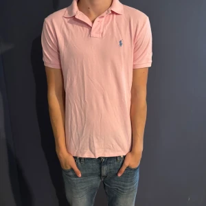 Rosa pikétröja Polo Ralph Lauren M - Snygg rosa pikétröja från Polo Ralph Lauren i modellen Custom Slim Fit.  Tröjan är köpt i usa för ungefär 799kr. Mitt pris: 499kr eftersom tröjan fortfarande är bra skick och inte har några defekter. Hör av er vid frågor eller funderingar