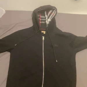 Svart Burberry hoodie med dragkedja - Svart hoodie från Burberry med klassiskt rutigt foder i huvan. Tröjan har hel dragkedja framtill, snörning i huvan och långa ärmar. Perfekt för dig som vill ha en stilren och lyxig streetwear-look.