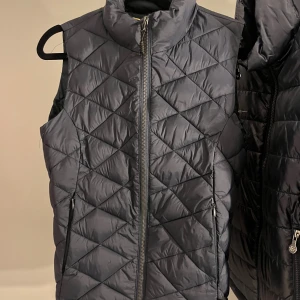 Mörkblå quiltad dunväst JH Collection XS - Snygg mörkblå dunväst från JH Collection i storlek XS. Västen är quiltad med ett mönster av stora rutor och har dragkedja framtill samt två fickor med dragkedja. Perfekt att slänga över en hoodie eller tröja för extra värme.
