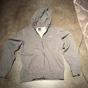 Grå Nike hoodie med dragkedja - Grå hoodie från Nike med vit broderad swoosh på bröstet. Tröjan har huva med snörning, hel dragkedja och två fickor framtill. Tillverkad i mjuk bomullsblandning, perfekt för chill eller träning.